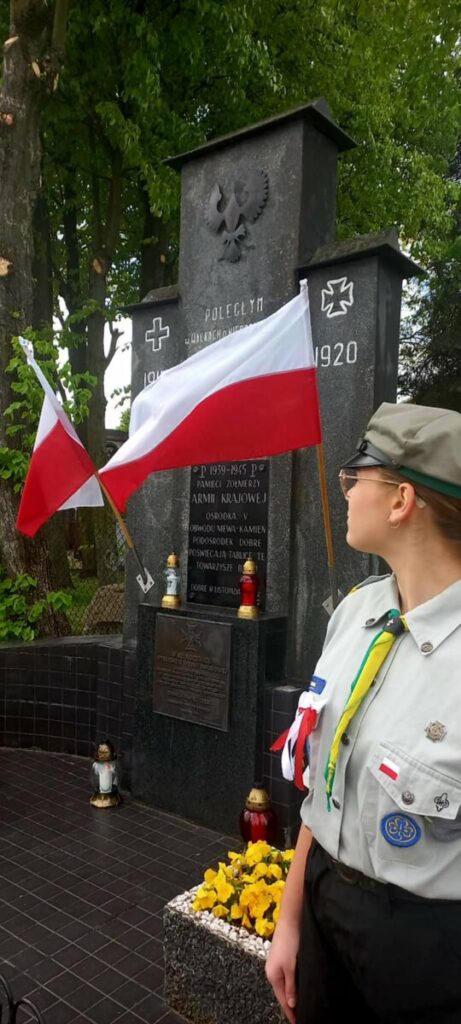Pomnik upamiętniający, otoczony drzewami, z biało-czerwonymi flagami. Harcerz w mundurze stoi przed nim. Kwiaty i znicze u podstawy.