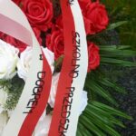 Bukiet czerwonych i białych róż z zielonymi liśćmi. Na wstążce czerwono-białej widnieje napis. Tło jest zielone i czarne.