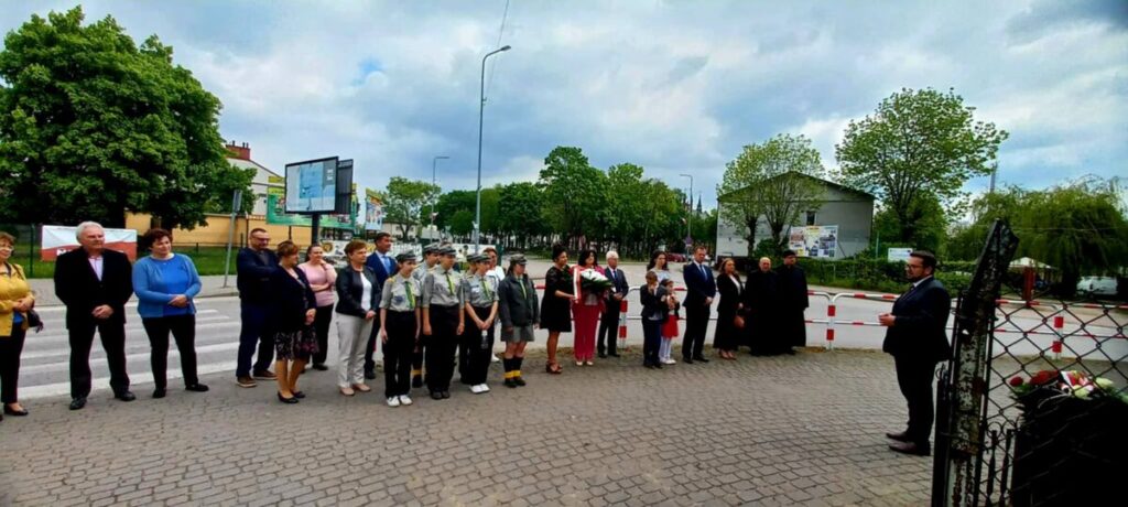 Ludzie stoją w szeregu na chodniku, jest pochmurne niebo. Harcerze w mundurach pośrodku. W tle drzewa i zabudowa miejska.