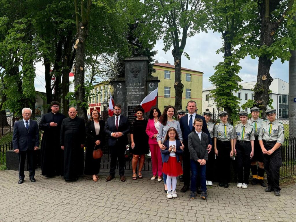 Grupa ludzi stoi przed pomnikiem w parku, w otoczeniu drzew. Mają formalne stroje, flagi Polski w tle. Tło to budynek i zielone drzewa.