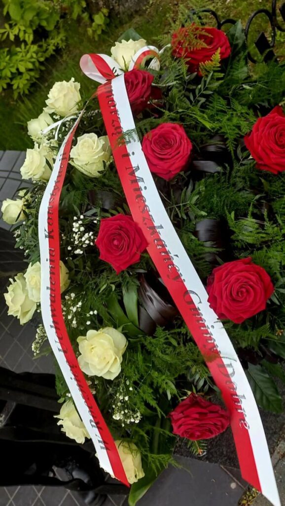 Wieniec z czerwonymi i białymi różami oraz zielonymi liśćmi. Na wieńcu biała wstęga z napisem. Tło to zieleń ogrodu.