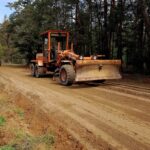 Pomarańczowa równiarka na ziemistej drodze w lesie. Tło pełne drzew i jasne niebo. Świeżo wyrównana powierzchnia.