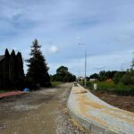 Droga w budowie z nowym, szarym chodnikiem z kostki brukowej. Po lewej stronie drzewa i krzewy, a w tle domy. Niebo jest jasnoniebieskie z niewielką ilością chmur.