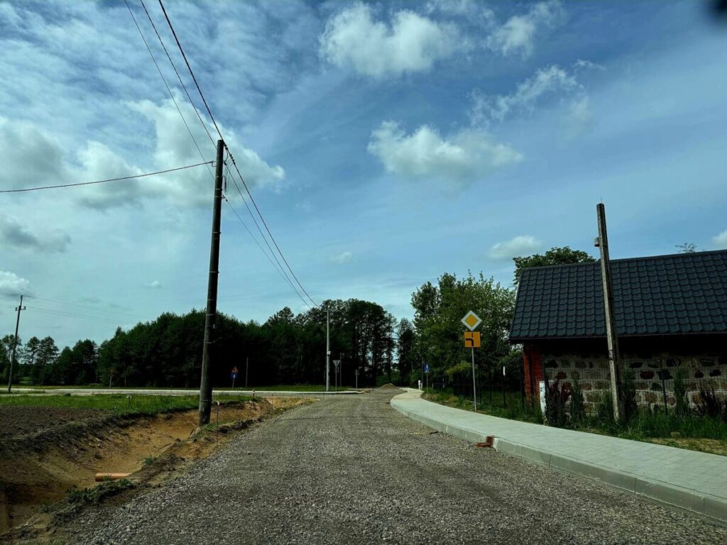 Droga żwirowa prowadzi obok budynku z kamienną ścianą i ciemnym dachem. Obok widoczny znak drogowy i zieleń drzew. Słoneczny dzień z niebieskim niebem i chmurami w tle.
