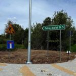 Znak drogowy wskazuje drogę do Zdrojówek, obok znak robót. Na tle drzewa, chodnik i lampa. Dzień jest pochmurny.