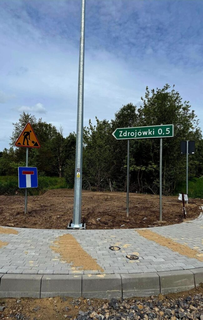 Znak drogowy wskazuje drogę do Zdrojówek, obok znak robót. Na tle drzewa, chodnik i lampa. Dzień jest pochmurny.