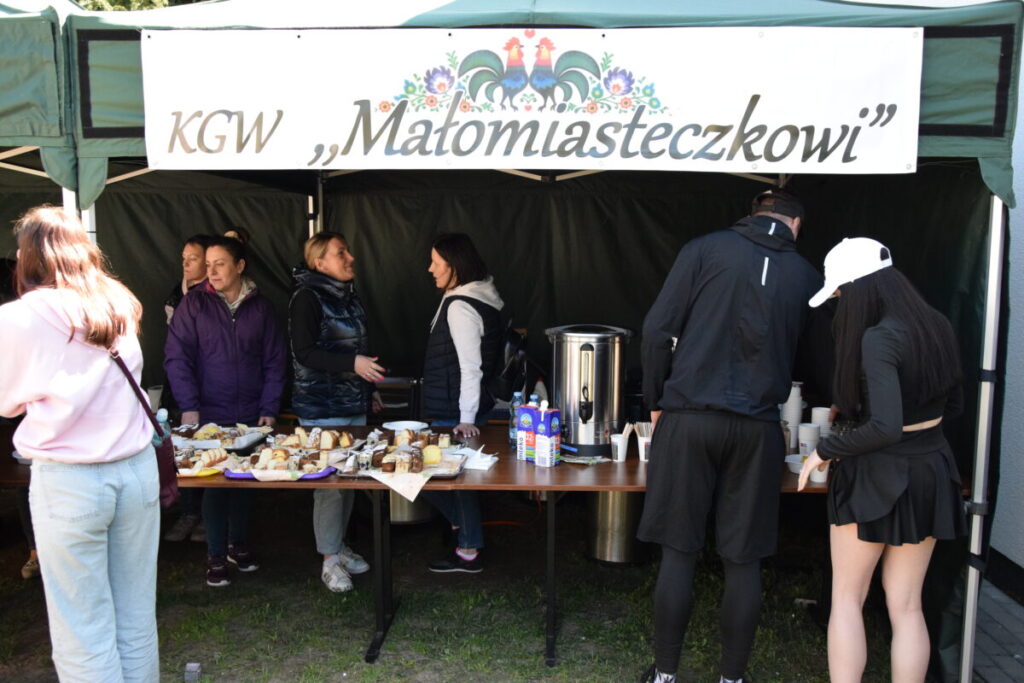 Stoisko KGW Małomiasteczkowi na tle zielonego namiotu. Na stole desery i napoje. Ludzie rozmawiają i częstują się.