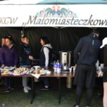 Stoisko KGW Małomiasteczkowi na tle zielonego namiotu. Na stole desery i napoje. Ludzie rozmawiają i częstują się.