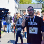 Na zdjęciu mężczyzna z mikrofonem w czarnej koszulce, otoczony uczestnikami wydarzenia na zewnątrz. Kamera w lewym rogu. Tło to jasny budynek i drzewa.