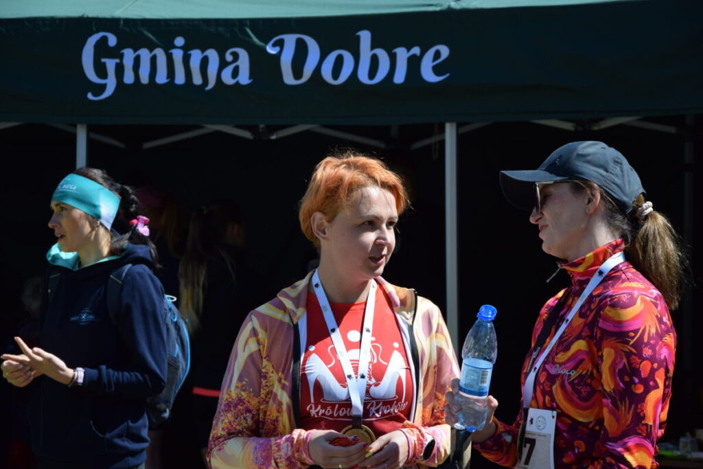 Trzy kobiety stoją przed namiotem z napisem Gmina Dobre. Jedna ma pomarańczową bluzę i medal, druga pije wodę. Kolory są jasne i żywe, tło ciemniejsze.
