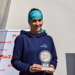 Osoba w niebieskiej bluzie z nagrodą, na tle białej ściany i banerów. Opaska turkusowa na głowie. Słońce świeci jasno.