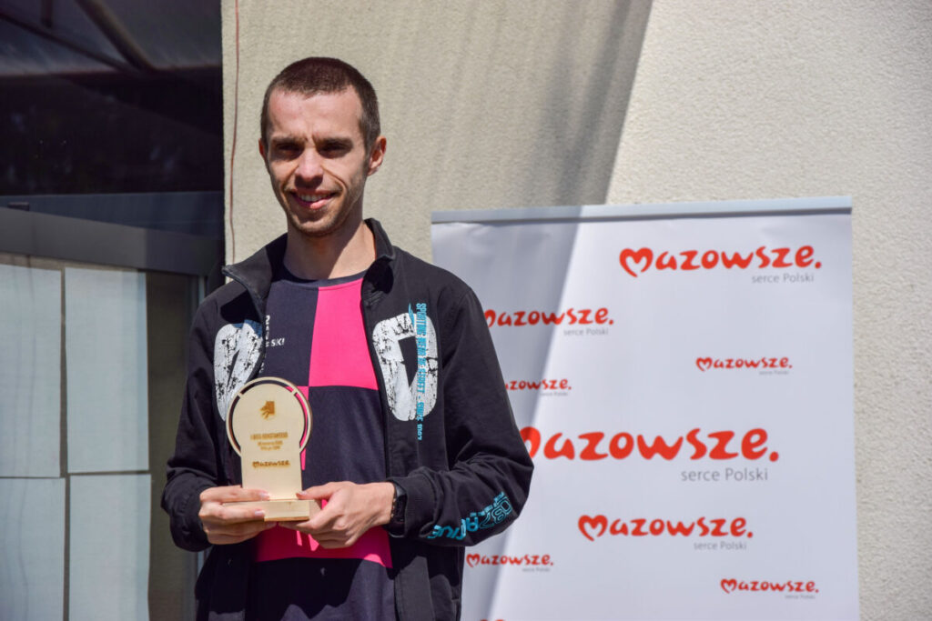 Osoba trzymająca trofeum stoi przed banerem „Mazowsze”. Ubrana w czarno-różowy strój, tło jasnobeżowe i biały baner.