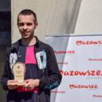 Osoba trzymająca trofeum stoi przed banerem „Mazowsze”. Ubrana w czarno-różowy strój, tło jasnobeżowe i biały baner.