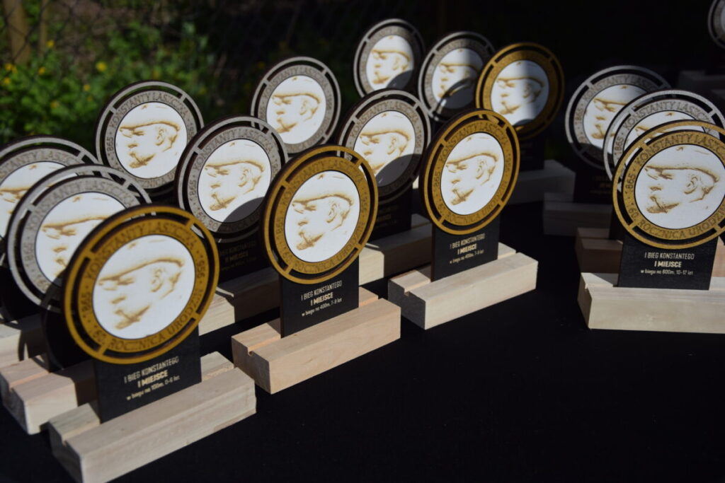 Rzędy okrągłych trofeów z profilową sylwetką na złotym i czarnym tle. Trofea stoją na jasnych drewnianych podstawach. W tle widoczna zieleń.