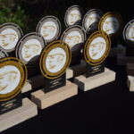 Rzędy okrągłych trofeów z profilową sylwetką na złotym i czarnym tle. Trofea stoją na jasnych drewnianych podstawach. W tle widoczna zieleń.