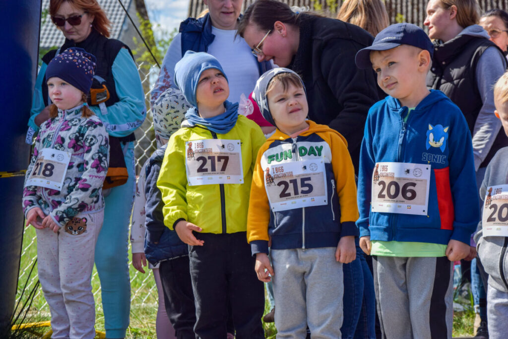 Grupa dzieci w kolorowych ubraniach stoi na świeżym powietrzu z numerami startowymi. Uśmiechnięte twarze, niebieskie i żółte kurtki. W tle widoczni dorośli i zielona trawa.