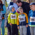 Grupa dzieci w kolorowych ubraniach stoi na świeżym powietrzu z numerami startowymi. Uśmiechnięte twarze, niebieskie i żółte kurtki. W tle widoczni dorośli i zielona trawa.