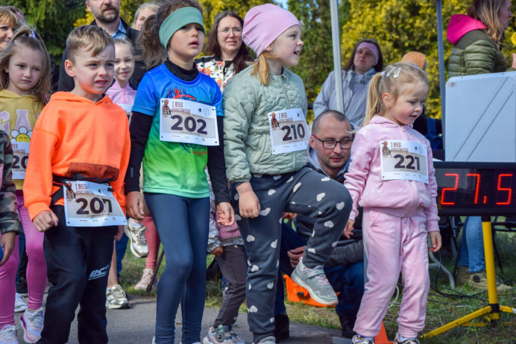 Na zdjęciu widać grupkę dzieci z numerami startowymi, przygotowujących się do biegu. Dzieci są ubrane w kolorowe stroje sportowe. W tle widoczni dorośli kibicują oraz wyświetlacz z czasem.