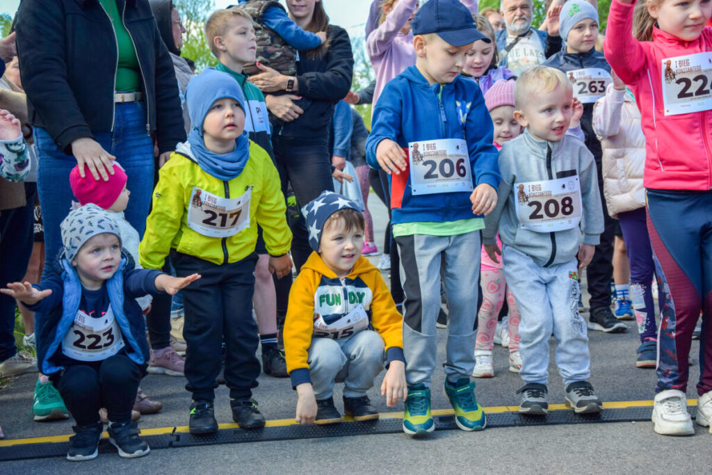 Grupa małych dzieci w kolorowych ubraniach stoi na linii startu. Mają numery startowe na torsach. W tle widoczni są dorośli i inne dzieci.