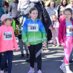 Grupa dzieci w kolorowych ubraniach i numerami startowymi na ulicy. Jasne słońce, wokół tłum ludzi. Tło jest rozproszone.