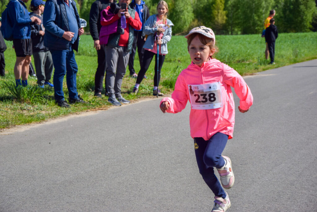 Dziewczynka biegnie w jasnoróżowej kurtce z numerem 238 po asfaltowej drodze. W tle soczysta zieleń trawy i drzewa. Na poboczu stoją kibice.