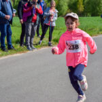 Dziewczynka biegnie w jasnoróżowej kurtce z numerem 238 po asfaltowej drodze. W tle soczysta zieleń trawy i drzewa. Na poboczu stoją kibice.