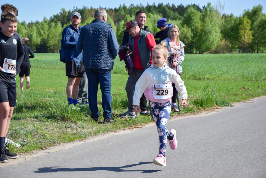 Dziewczynka biegnie w zawodach na asfaltowej drodze, ubrana kolorowo. W tle zielone pole i ludzie obserwujący. Ciepły, słoneczny dzień.