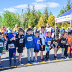 Grupa dzieci stoi na linii startu biegu. Są ubrani w sportowe stroje, z numerami na koszulkach. Tło zdobią drzewa i namiot.