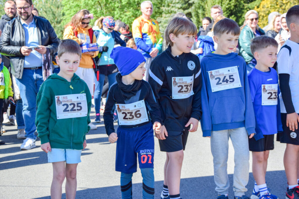 Grupa dzieci w kolorowych strojach sportowych z numerami startowymi stoi na drodze. Za nimi widoczny tłum dorosłych. Słoneczna pogoda, zieleń w tle.