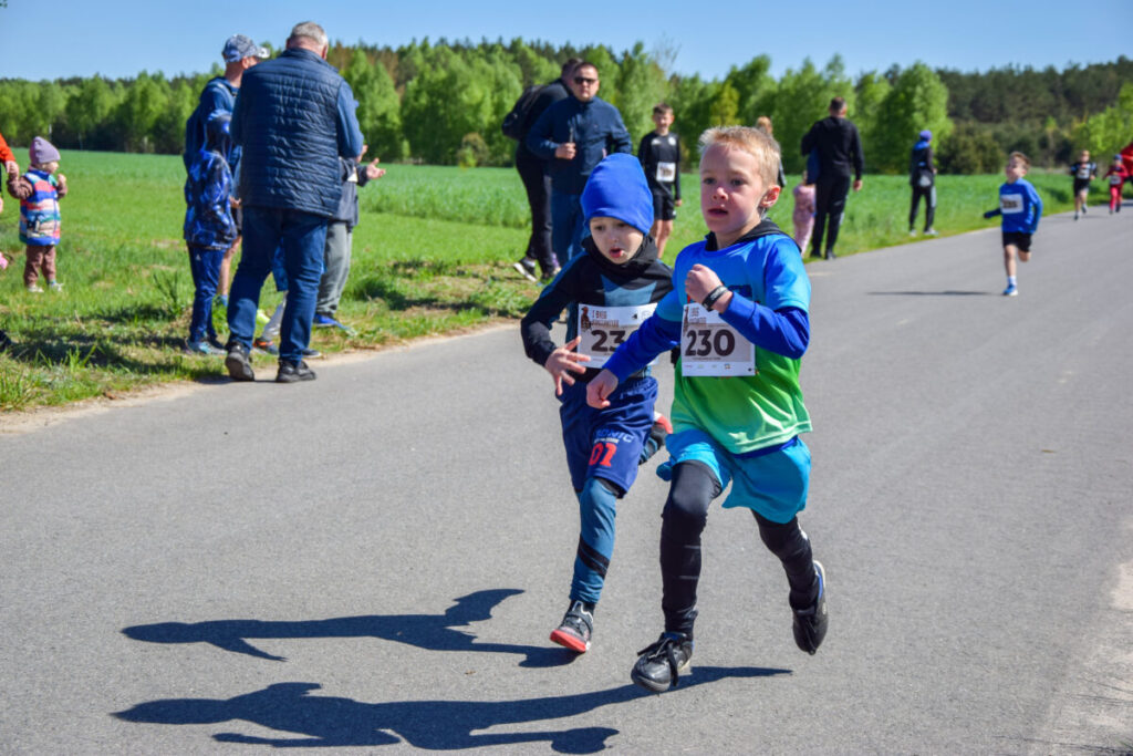 Dwaj chłopcy biegną w wyścigu na wiejskiej drodze, ubrani na niebiesko i zielono. Tło to zielone pola i drzewa. Towarzyszy im grupa ludzi obserwujących z boku.