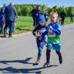 Dwaj chłopcy biegną w wyścigu na wiejskiej drodze, ubrani na niebiesko i zielono. Tło to zielone pola i drzewa. Towarzyszy im grupa ludzi obserwujących z boku.