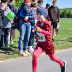 Dziecko w czerwonym stroju sportowym biegnie po asfalcie. W tle kibicujący ludzie i zielone pole. Słoneczna pogoda, atmosfera sportowa.