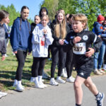 Chłopiec w czarnym stroju biegnie z numerem 237. Kibice w bluzach stoją obok, a w tle widoczna zieleń drzew. Dzień jest słoneczny.