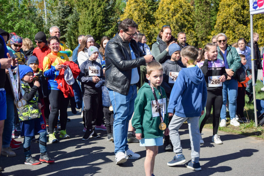 Grupa ludzi, w tym dzieci, uczestniczy w biegu na świeżym powietrzu. Tło z zielonymi drzewami. Jasne, słoneczne kolory.