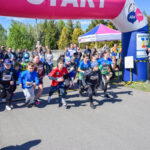Na zdjęciu dzieci rozpoczynają bieg spod różowego banera START. Ubrane są kolorowo, a w tle widać zielone drzewa. Wokół stoją klaskający widzowie.