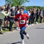 Chłopiec w czerwonej koszulce biegnie podczas zawodów. W tle grupa kibiców na zielonej polanie. Słoneczny dzień.