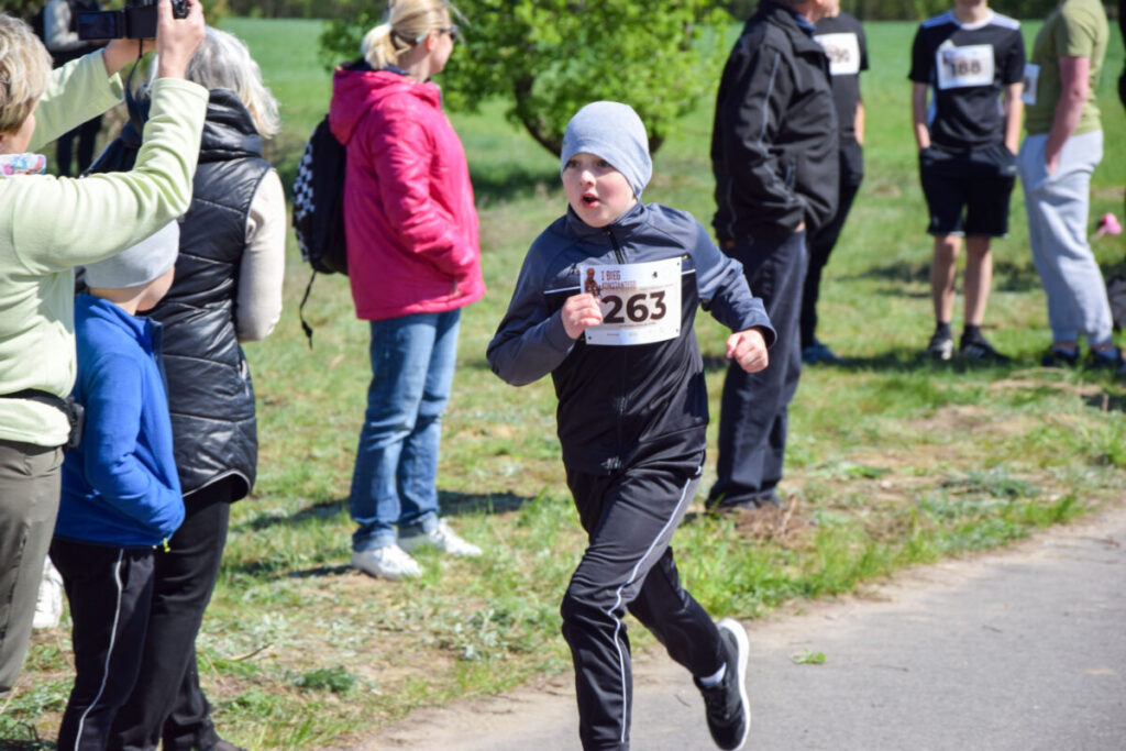 Chłopiec w czapce biegnie podczas wyścigu na świeżym powietrzu. Nosi czarny strój, numer startowy 263. W tle trawa i stojący ludzie.