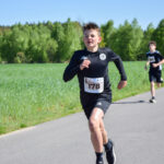 Chłopiec w czarnym stroju biegowym z numerem startowym 178 biegnie po asfaltowej ścieżce. W tle zielone pola i drzewa. Słoneczna pogoda, jasne niebo.