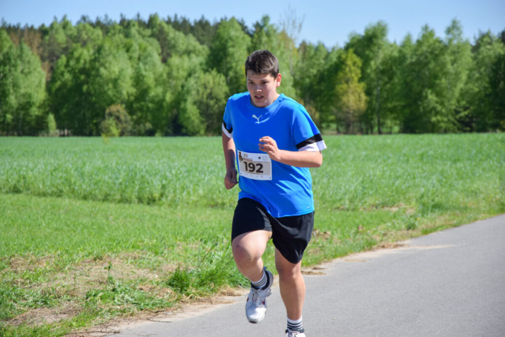 Na zdjęciu chłopiec biegnie w stroju sportowym z numerem 192. Ma niebieską koszulkę, a za nim jest zielone pole i las. Jest słoneczny dzień z błękitnym niebem.