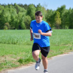 Na zdjęciu chłopiec biegnie w stroju sportowym z numerem 192. Ma niebieską koszulkę, a za nim jest zielone pole i las. Jest słoneczny dzień z błękitnym niebem.