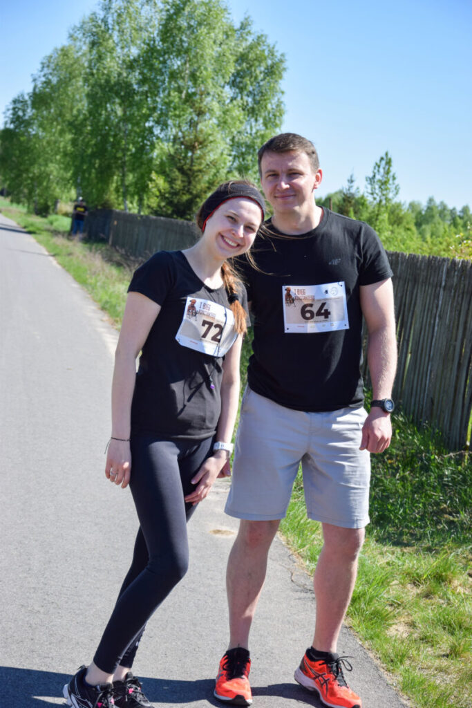 Para w sportowych strojach z numerami startowymi pozuje na asfaltowej drodze. Tło stanowią zielone drzewa i drewniany płot. Jasne niebo podkreśla słoneczną pogodę.