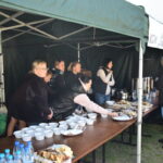 Grupa ludzi obsługuje stoisko z jedzeniem pod zielonym namiotem. Na stole są talerze z przekąskami i napoje. Tło jest w szarym odcieniu.