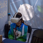 Osoba w niebieskiej kurtce nosi gogle VR, siedząc przy stole. Tło to niebieski baner z godłem Polski. Obok jest biały kubek.