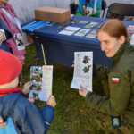 Na zdjęciu żołnierka w zielonym mundurze pokazuje chłopcu broszury. Chłopiec ma czerwony kaptur. W tle stół z materiałami promocyjnymi.