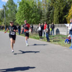 Dwóch biegaczy w trakcie wyścigu na asfaltowej drodze. Wokół kibicują widzowie na zielonym tle drzew. Słońce rzuca cienie.