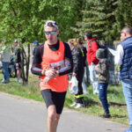 Mężczyzna biegnie w wyścigu, ubrany w pomarańczową kamizelkę. Tło to zielone drzewa i kibice. Dzień jest jasny i słoneczny.