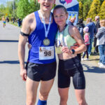 Para biegaczy z medalami uśmiecha się przed bramką startową. Niebo jest niebieskie, a w tle widać zielone drzewa i ludzi.