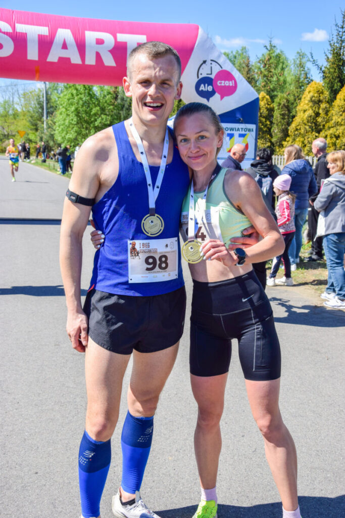Para biegaczy z medalami uśmiecha się przed bramką startową. Niebo jest niebieskie, a w tle widać zielone drzewa i ludzi.
