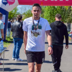 Na zdjęciu widać biegacza w białej koszulce i czarnych szortach, przechodzącego pod różowym balonem z napisem START. Tło stanowi zielona roślinność i niebo. Obok biegacza stoi stół z wodą dla uczestników.