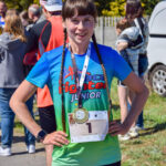 Kobieta w kolorowej koszulce z numerem startowym stoi po biegu. Ma medale na szyi. W tle inni uczestnicy i drzewa.